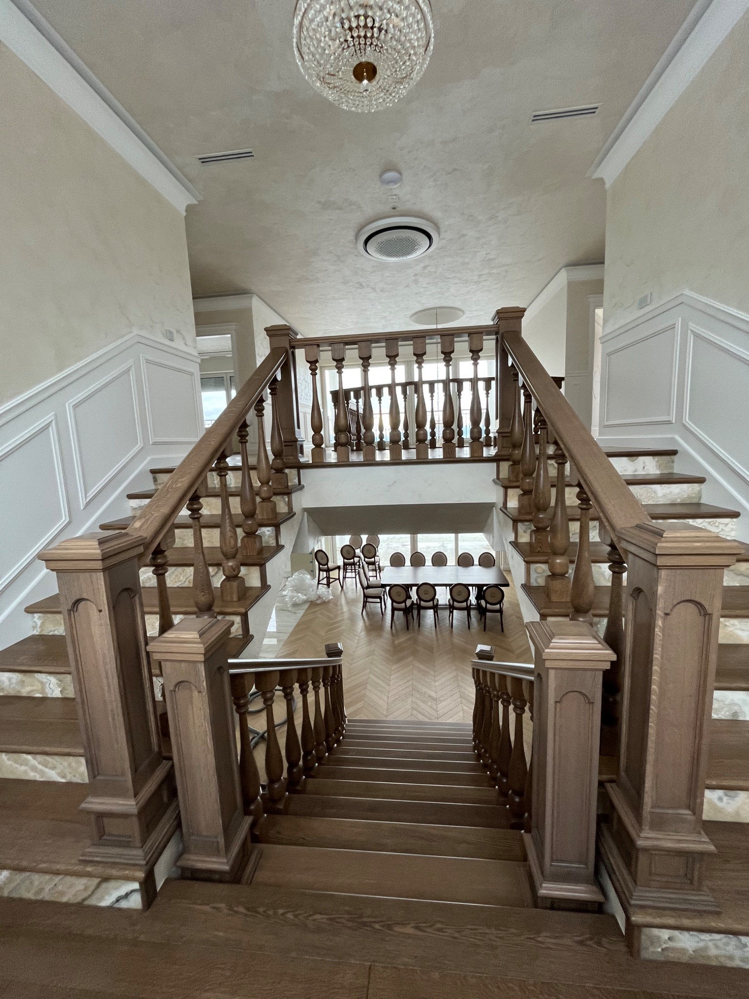 Dwubiegowe drewniane schody z balustradą i marmurowymi wstawkami na stopniach, widok z góry na dół, w tle parkiet ułożony w jodełkę i stół z krzesłami.