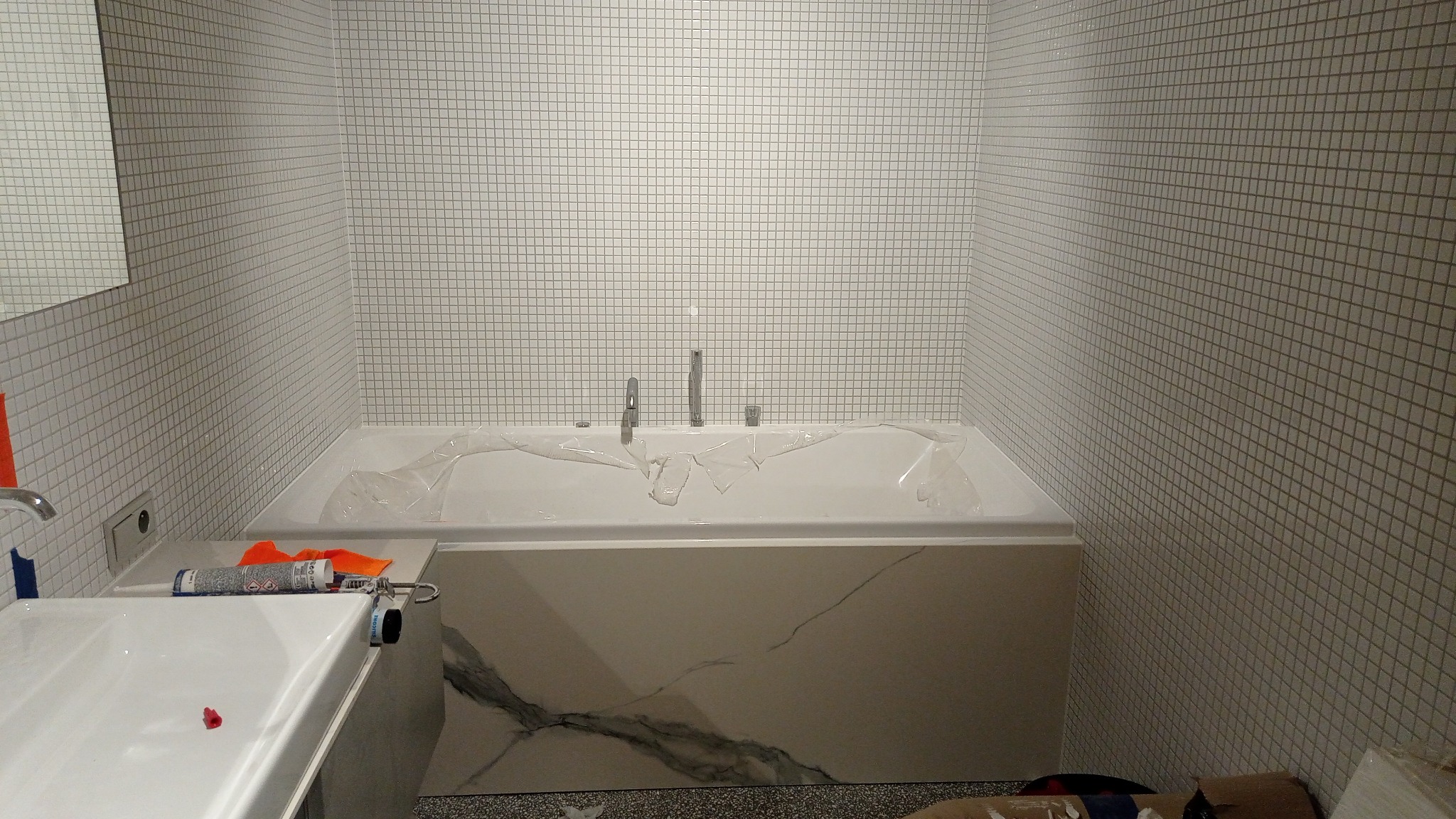 Biała wanna obłożona płytkami mozaikowymi, zabezpieczona folią, z widoczną baterią. Na blacie obok wanny leży tuba silikonu i pistolet do silikonu. Wykończenie łazienki.