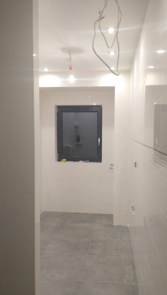 Wnętrze łazienki w trakcie wykańczania: szare płytki podłogowe, białe ściany z oknem. Widoczne niedokończone instalacje elektryczne i punktowe oświetlenie sufitowe.