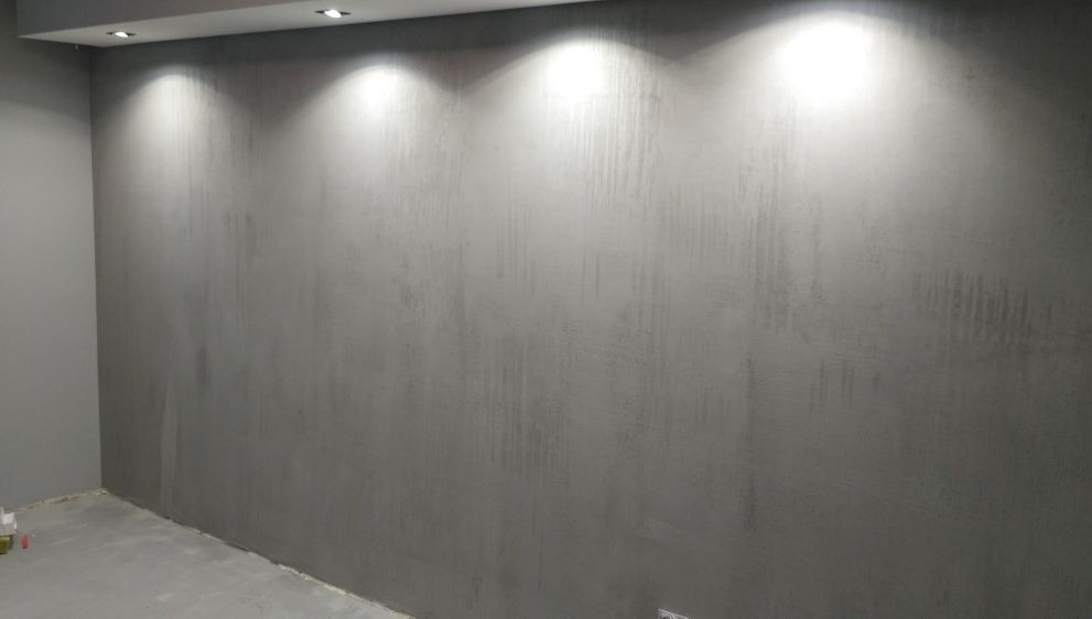 Szara ściana z efektem dekoracyjnym podświetlona trzema punktowymi lampami sufitowymi. Widoczny fragment podłogi i narożnik pokoju.