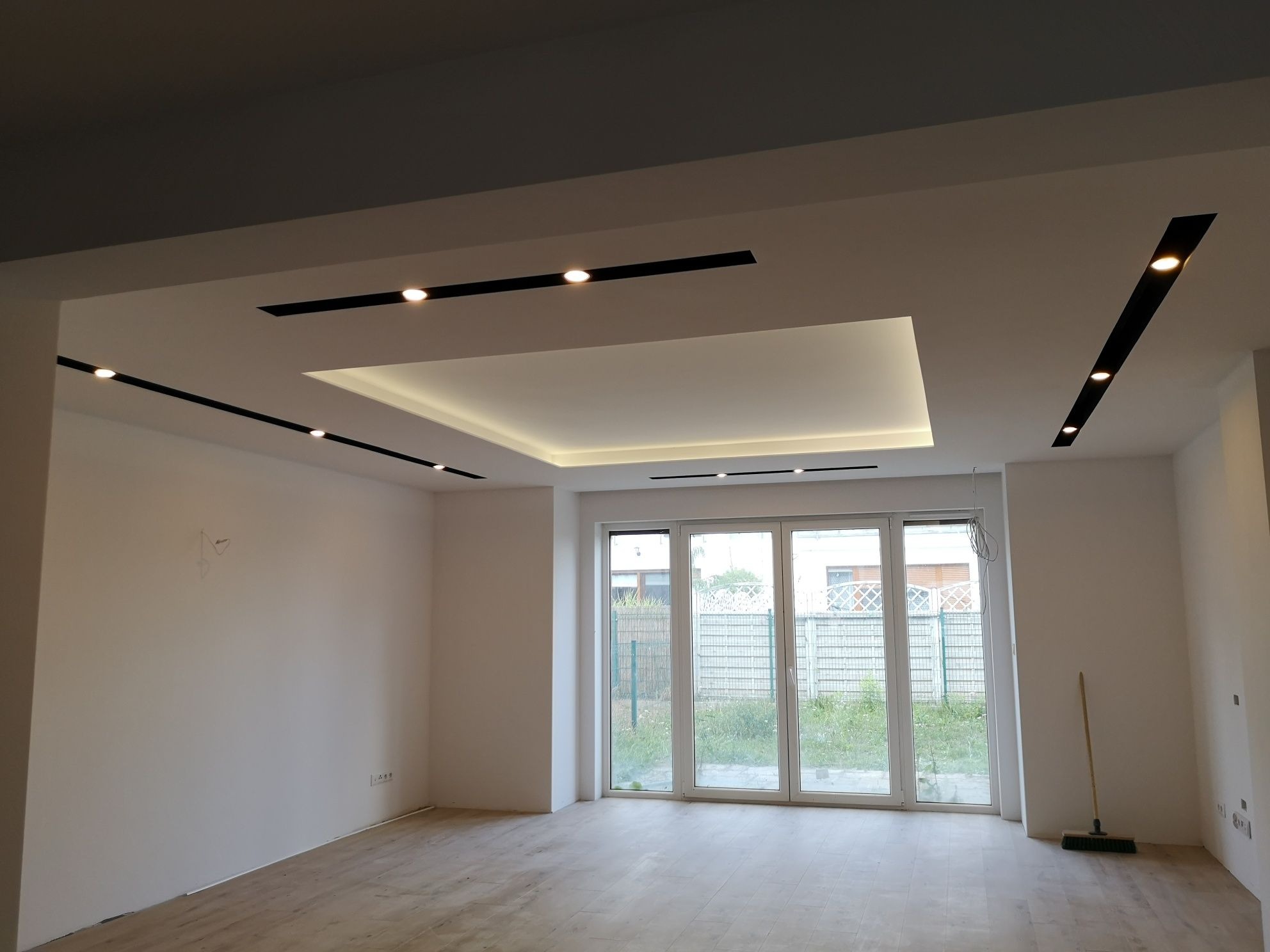 Nowoczesne wykończenie wnętrza: białe ściany, drewniana podłoga i sufit podwieszany z oświetleniem punktowym. Duże okno balkonowe z widokiem na zieleń.