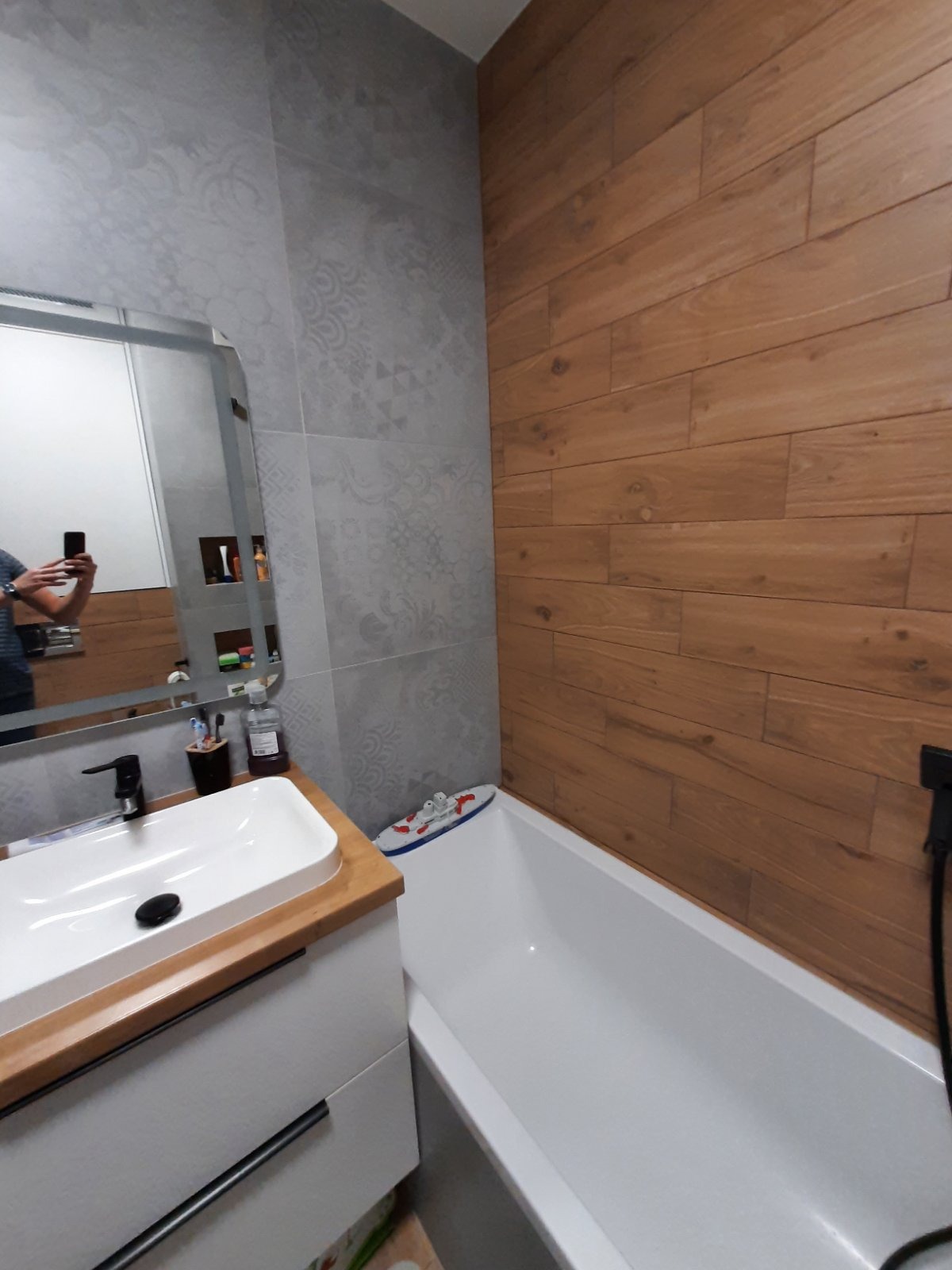 Nowoczesna łazienka po remoncie: drewnopodobne płytki, szara mozaika, biała wanna i umywalka z drewnianym blatem. W lustrze odbija się osoba robiąca zdjęcie.