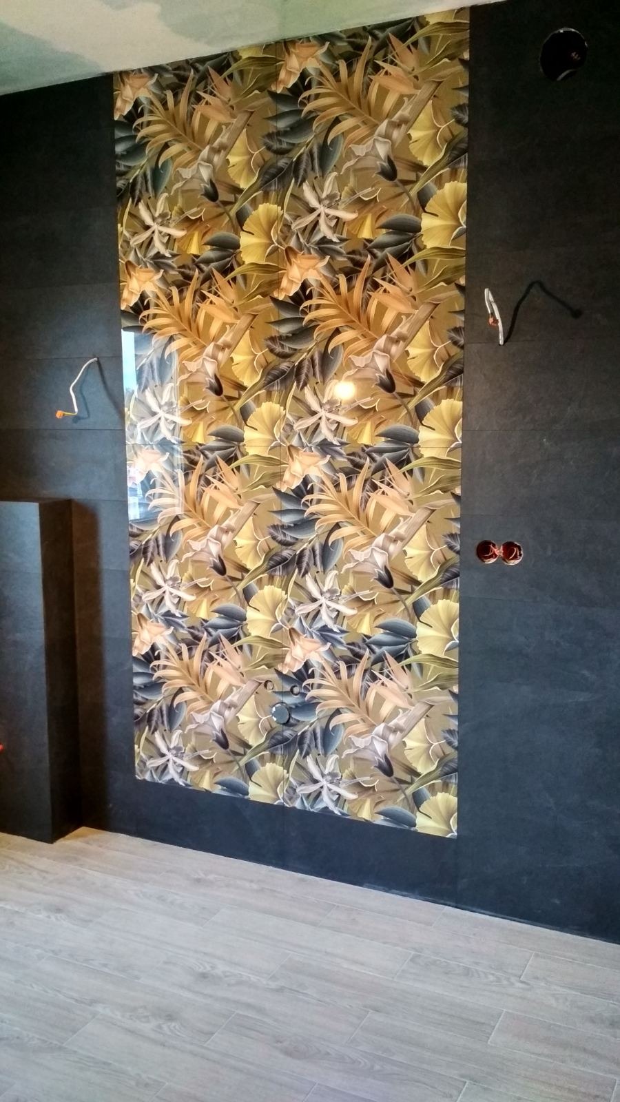 Wykończona ściana z wzorzystymi, błyszczącymi płytkami w motywy roślinne, sąsiadująca z ciemną ścianą z widocznymi gniazdkami i fragmentem instalacji elektrycznej.