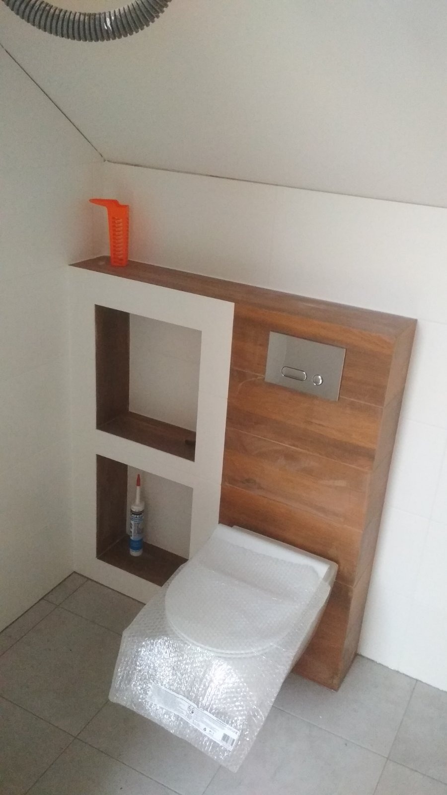 Wisząca toaleta w folii ochronnej, zabudowana drewnianą płytą z przyciskiem spłukiwania. Nad nią wnęka z półkami, w której stoją akcesoria montażowe.