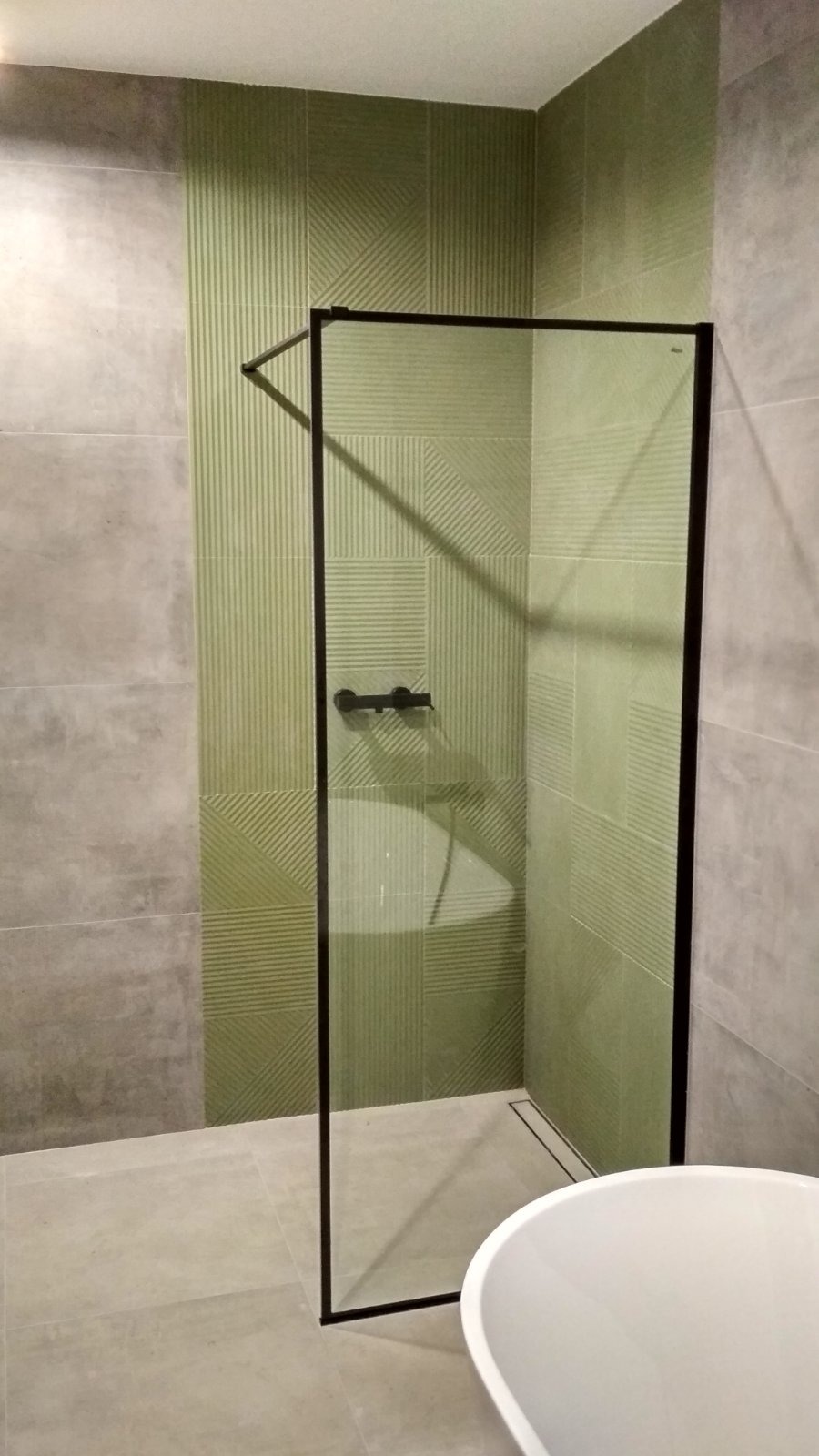 Nowoczesna kabina prysznicowa z czarną ramą i zielonymi płytkami o geometrycznym wzorze. Minimalistyczny design łazienki, widoczny fragment wanny.