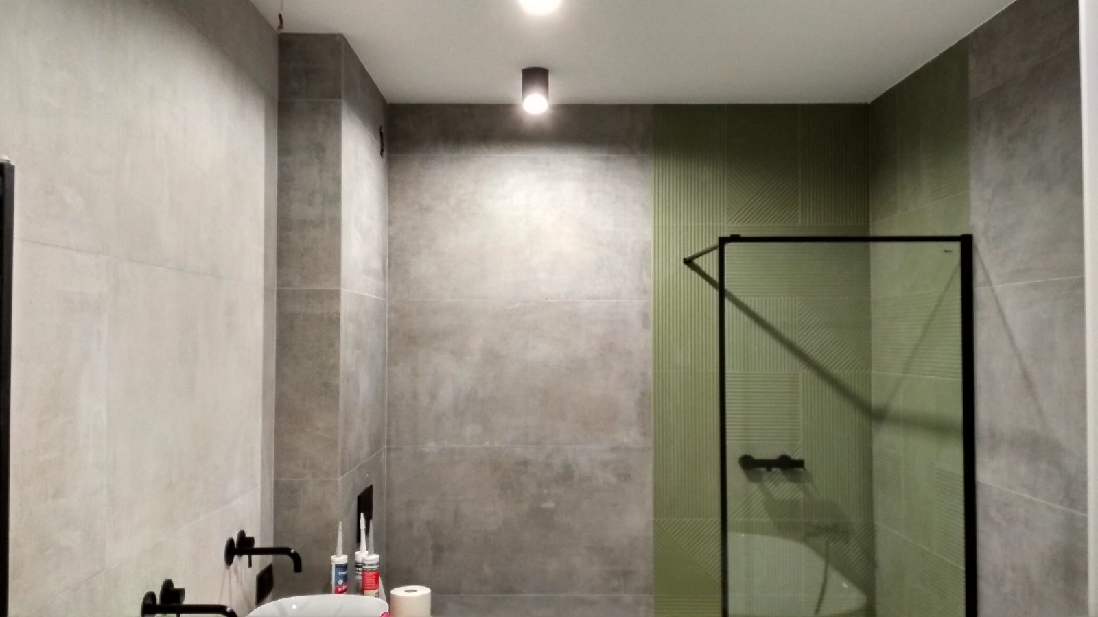 Wykończona łazienka z kabiną prysznicową z czarnymi ramami i zielonymi płytkami. Na ścianach szare płytki imitujące beton. Widoczne czarne baterie umywalkowe.