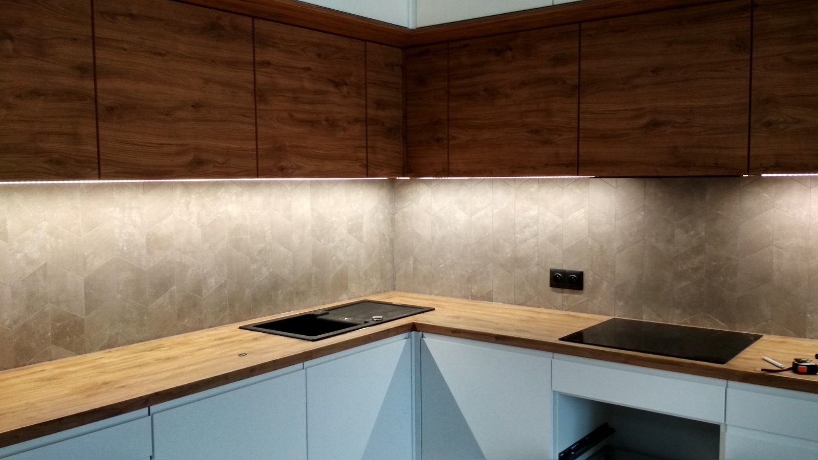 Nowoczesna kuchnia z drewnianymi szafkami, blatem i heksagonalnymi płytkami. Podświetlenie LED dodaje ciepła. Widoczny zlew i płyta indukcyjna.