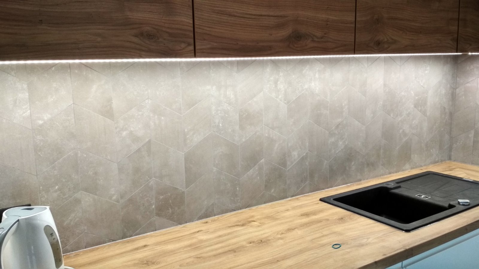 Wykończona ściana w kuchni z szarymi, heksagonalnymi płytkami podświetlonymi listwą LED, drewniany blat z czarnym zlewem. Nowoczesny design.