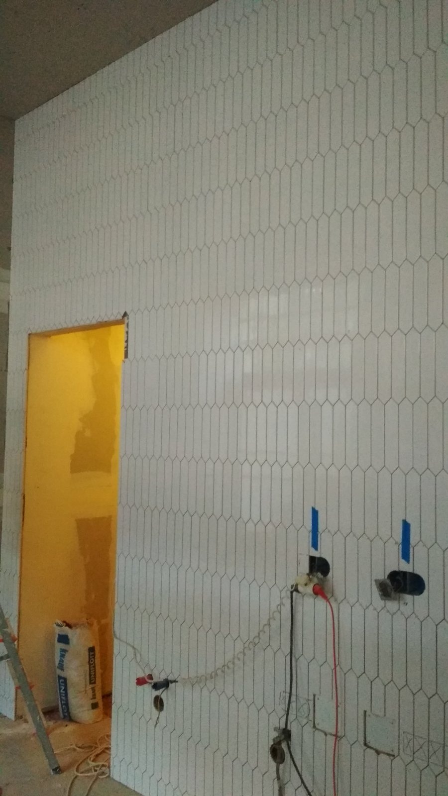 Ściana wyłożona białymi, podłużnymi płytkami w kształcie rombów. Widoczne wyprowadzenia instalacji elektrycznej. W tle torba z zaprawą i fragment drabiny.