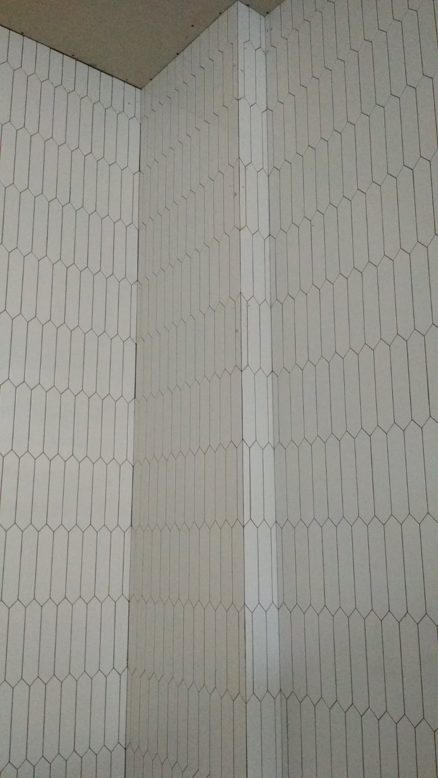 Białe, podłużne płytki ułożone pionowo w geometryczny wzór na ścianie w rogu pomieszczenia, widok z dołu.