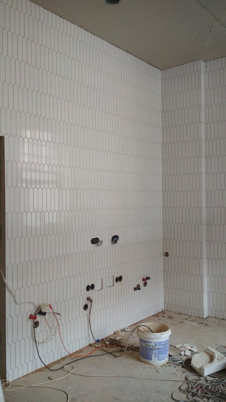 Ściana wyłożona białymi, pionowymi płytkami o kształcie wydłużonego owalu. Widoczne otwory na instalacje. Na podłodze resztki materiałów budowlanych i wiadro.