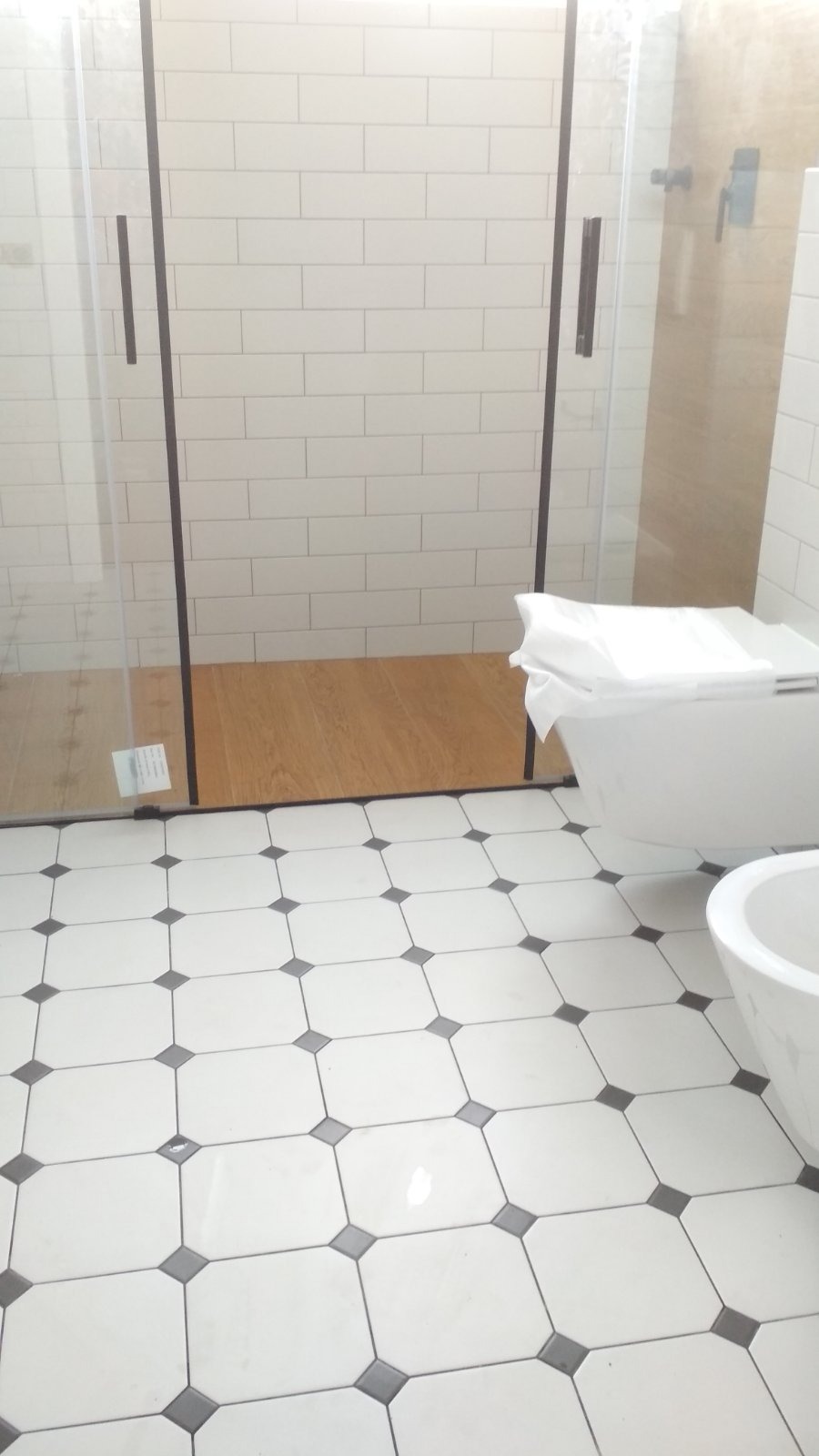 Wykończona łazienka z kabiną prysznicową typu walk-in, białą ceramiką i podłogą w biało-czarną szachownicę. Widoczne białe, prostokątne płytki na ścianach.