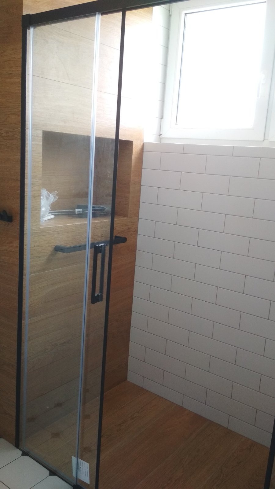 Nowoczesna kabina prysznicowa z przesuwnymi drzwiami, wykończona płytkami imitującymi drewno i białymi cegiełkami. Wnęka z półką na akcesoria.