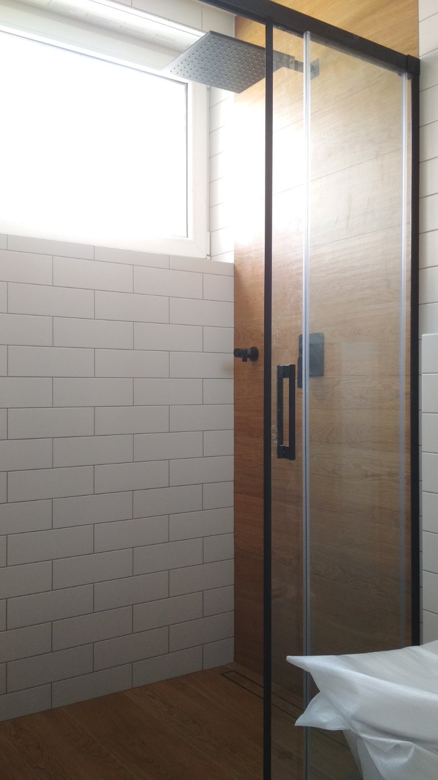 Nowoczesna kabina prysznicowa z deszczownicą i drewnianym panelem, białe kafelki w stylu metro, czarne detale armatury. Minimalistyczny design łazienki.