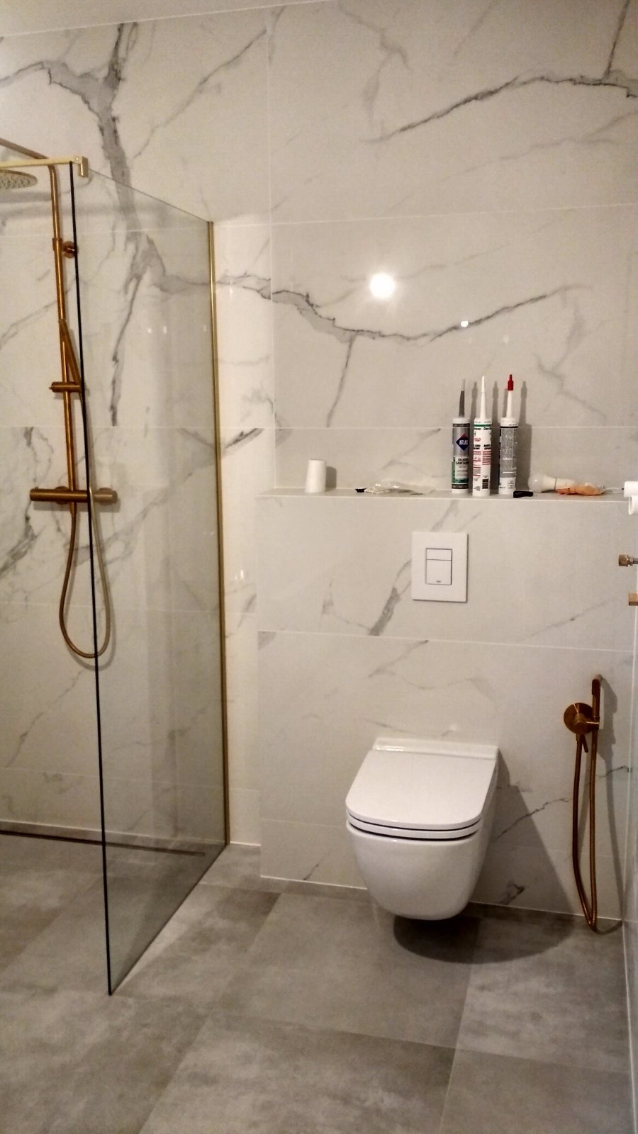 Nowoczesna łazienka z marmurowymi płytkami, złotą armaturą prysznicową i podwieszaną toaletą. Widoczne tuby silikonu na półce.