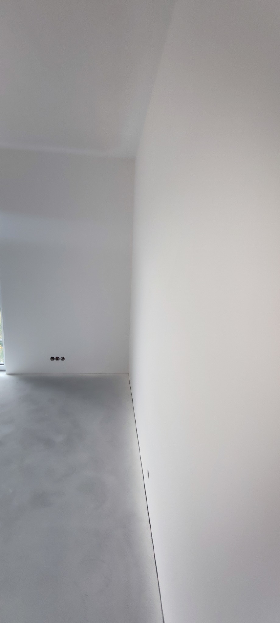 Białe, gładkie ściany po szpachlowaniu w pustym pokoju z betonową podłogą i widocznymi gniazdkami elektrycznymi. Minimalistyczne wnętrze w trakcie wykańczania.