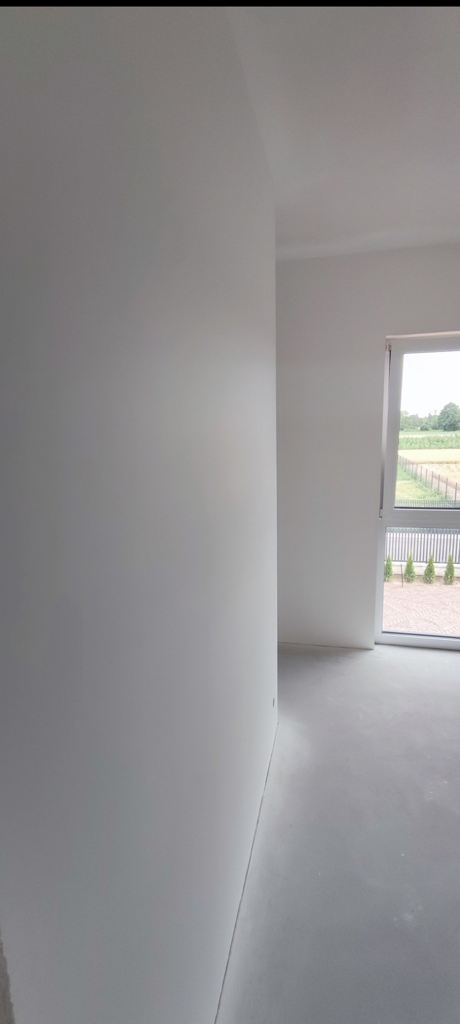 Jasne, świeżo pomalowane wnętrze z oknem, ukazujące gładkie, białe ściany i podłogę. Widok na zewnątrz przez okno z zielenią w tle.