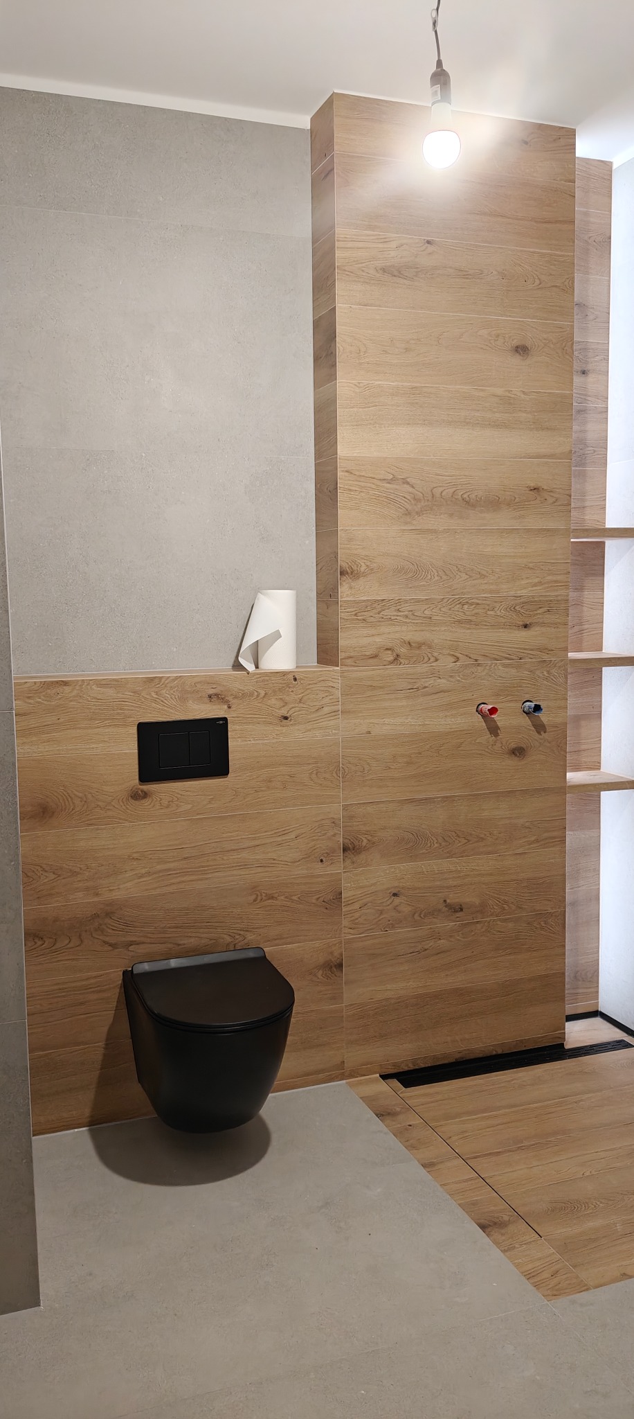 Nowoczesna łazienka z drewnopodobnymi płytkami, wiszącą toaletą i szarą podłogą. Minimalistyczny design, widoczne detale instalacji wodnej i oświetlenie.