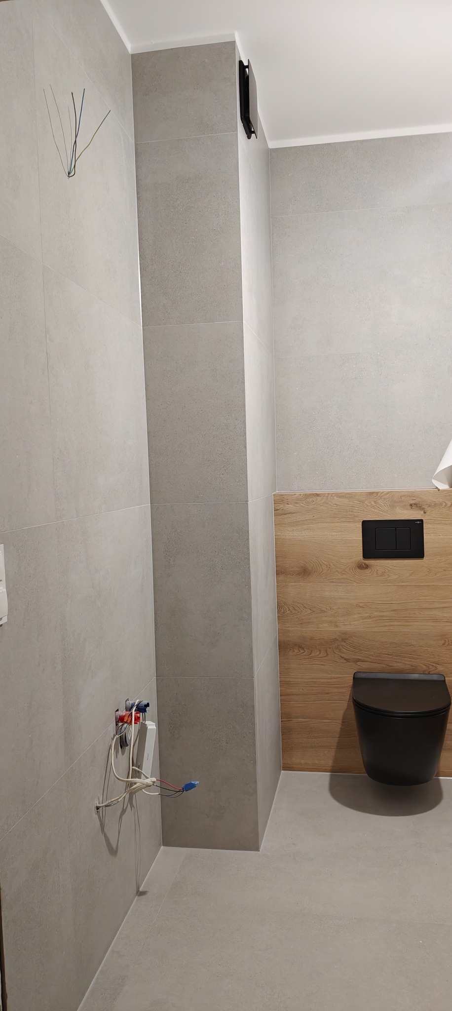 Wykończona łazienka z szarymi płytkami, drewnopodobną zabudową i podwieszaną toaletą. Widoczne wystające kable i rurki przed montażem armatury. Minimalistyczny design.
