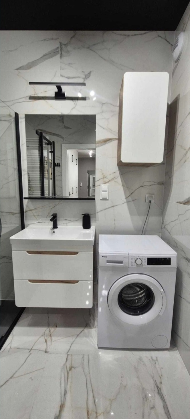 Nowoczesna łazienka z marmurowymi płytkami, białą szafką pod umywalką, pralką Amica i lustrem z czarną ramą. Widoczny fragment kabiny prysznicowej.