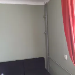 Fragment pokoju z pionową rurą hydrauliczną przy ścianie pomalowanej na oliwkowy kolor, widoczny fragment sofy i czerwona zasłona.