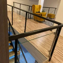 Szklana balustrada z czarnymi metalowymi słupkami, widok z góry na drewniane schody i podłogę, w tle żółty fotel uszak i pufa w jasnym wnętrzu.