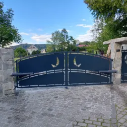 Automatyczna brama wjazdowa dwuskrzydłowa z dekoracyjnymi wycięciami w kształcie półksiężyców, zamontowana na kamiennych słupkach, z widocznymi siłownikami i kostką brukową na podjeździe.