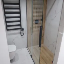 Nowoczesna łazienka z kabiną prysznicową typu walk-in, grzejnikiem drabinkowym w kolorze czarnym, umywalką wiszącą i płytkami imitującymi marmur na ścianach i podłodze, oraz płytkami imitującymi...