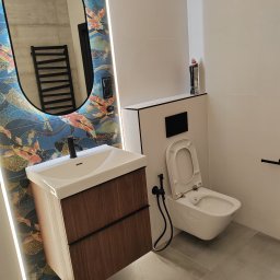 Budownictwo Komercyjne Grzegorz Bierecki - Nowoczesna łazienka z umywalką na drewnianej szafce, toaletą podwieszaną z bidetką i owalnym lustrem podświetlonym LED na ścianie z tapetą w orientalne wzory.