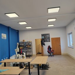 Budownictwo Komercyjne Grzegorz Bierecki - Przestronne, jasne wnętrze biura z niebieską ścianą, wyposażone w robotyczne ramię KUKA, biurka z czarnymi nogami, krzesło gamingowe i równomiernie rozmieszczone kwadratowe lampy sufitowe.