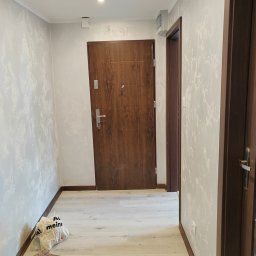 Budownictwo Komercyjne Grzegorz Bierecki - Korytarz po remoncie z drewnianymi drzwiami, panelami podłogowymi i ścianami z efektem dekoracyjnym, widoczne torby po zakupach przy ścianie.