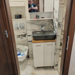 Budownictwo Komercyjne Grzegorz Bierecki - Wykończona łazienka z marmurowymi płytkami, szarą umywalką na drewnianym blacie i białymi szafkami pod umywalką, widok przez futrynę drzwi z odbiciem fotografa w lustrze.