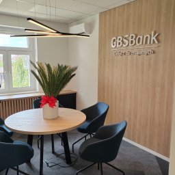 Budownictwo Komercyjne Grzegorz Bierecki - Jasne biuro z okrągłym stołem, czterema granatowymi krzesłami, rośliną w białej doniczce z czerwoną kokardą, nowoczesną lampą sufitową i ścianą z drewnianym panelem z logo GBS Bank.