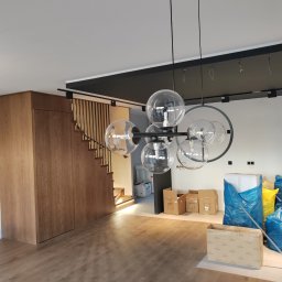 Budownictwo Komercyjne Grzegorz Bierecki - Nowoczesny żyrandol z kulistymi kloszami w jasnym wnętrzu z drewnianą podłogą i schodami, widoczne kartony i worki budowlane sugerujące prace wykończeniowe.