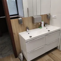 Nowoczesna łazienka z podwójną umywalką na szafce z szufladami, drewnianą podłogą i białymi płytkami. Widoczny dozownik mydła i okno z żaluzjami.