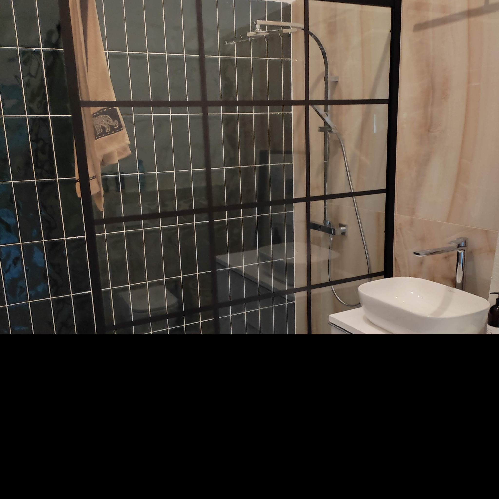 Nowoczesna łazienka z kabiną prysznicową typu walk-in z czarnymi profilami, umywalką nablatową i baterią w minimalistycznym stylu, na ścianach płytki w odcieniach zieleni i beżu.