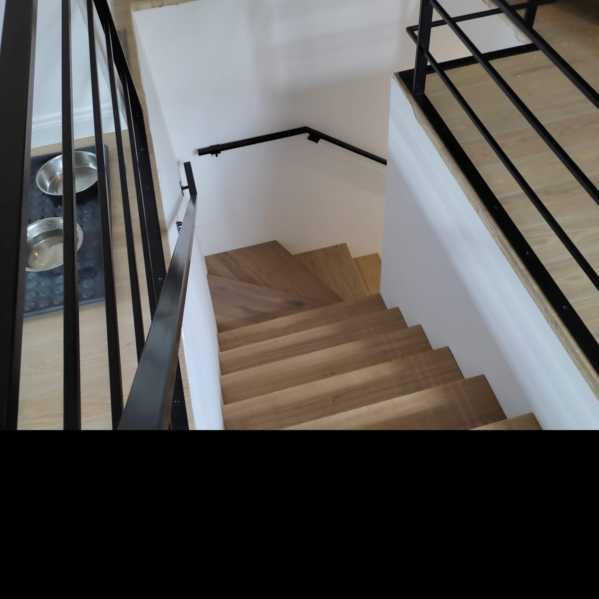 Drewniane schody z czarną, metalową balustradą, widok z góry, obok miski dla zwierząt na podłodze.