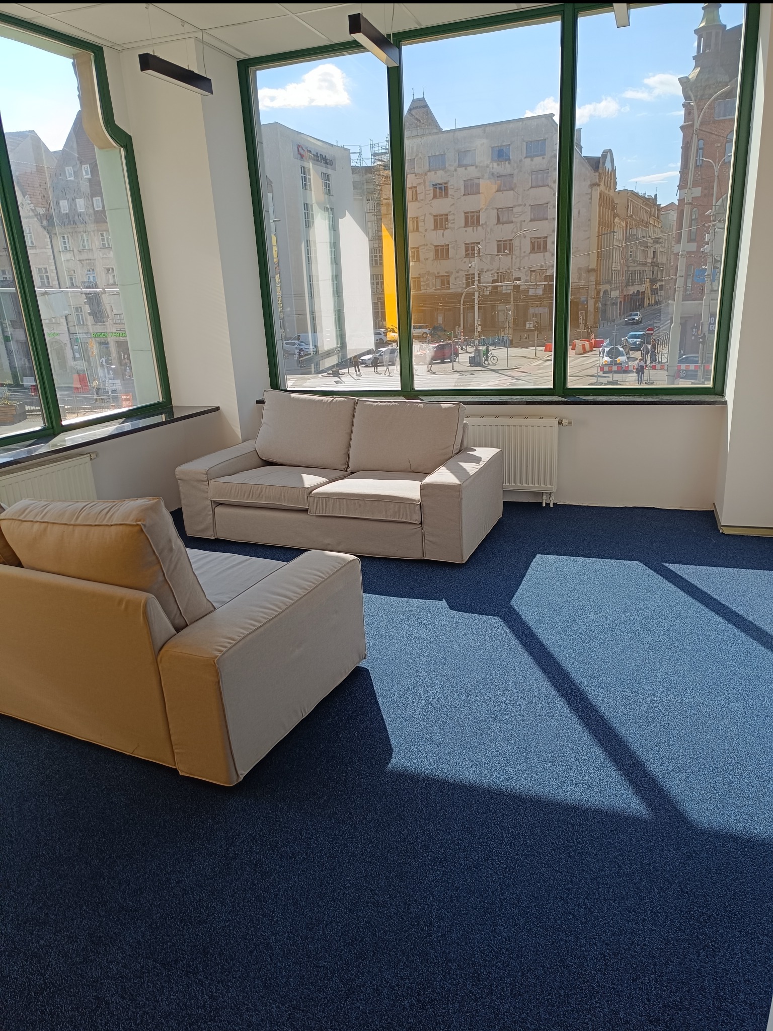 Jasne sofy w przestronnym biurze z widokiem na miasto przez duże okna z zielonymi ramami, niebieska wykładzina podłogowa.