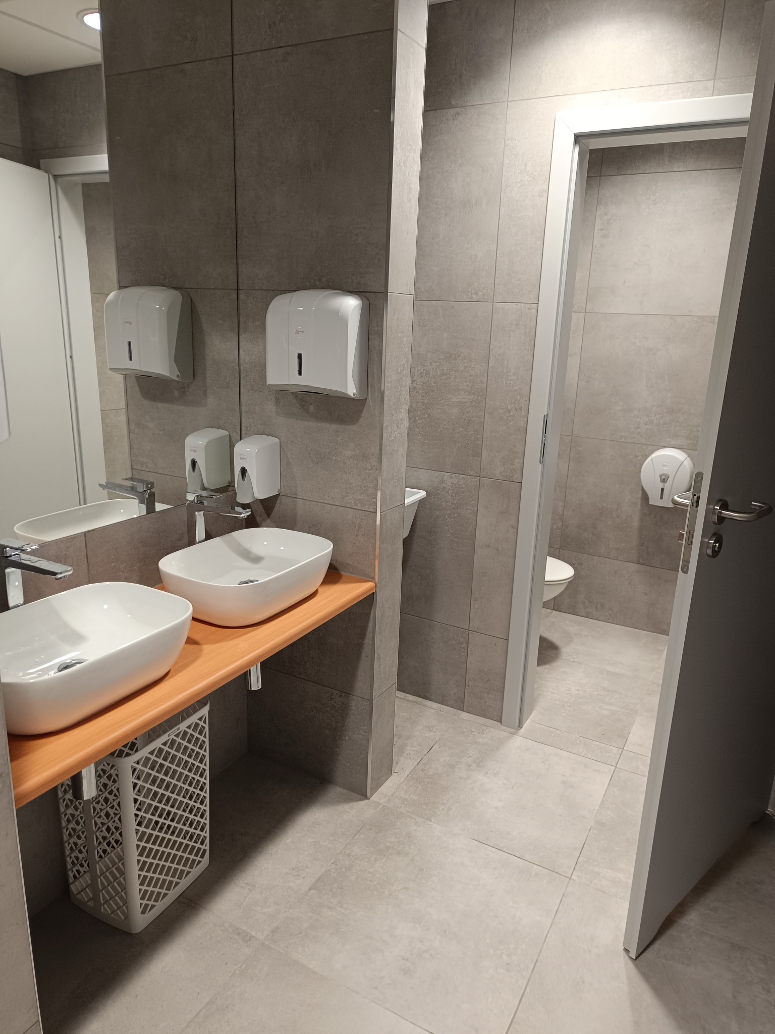 Nowoczesna łazienka z dwoma białymi umywalkami nablatowymi na drewnianym blacie, szare płytki na ścianach i podłodze, oraz widoczną toaletą w oddali.