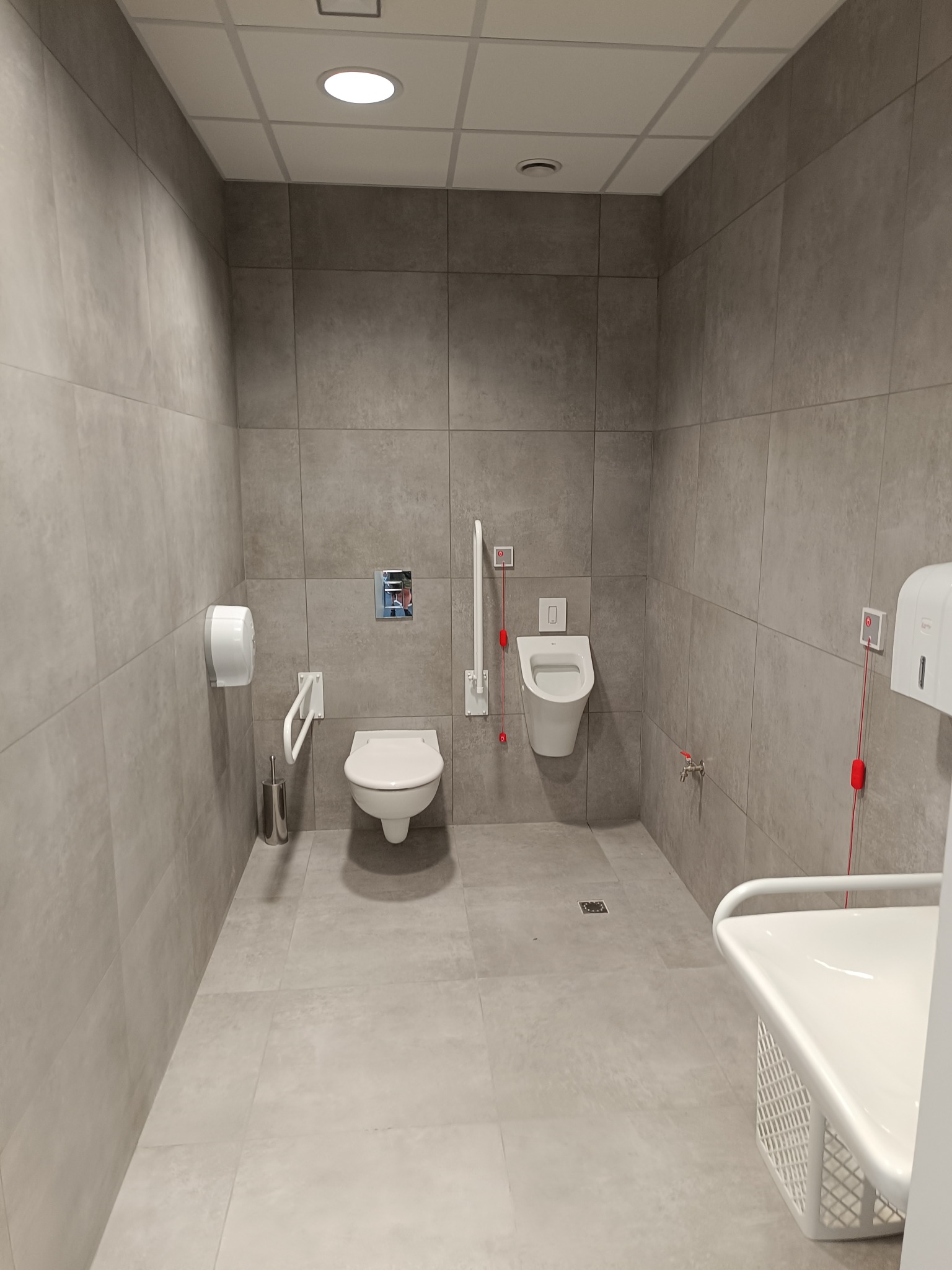 Jasna, minimalistyczna łazienka publiczna z udogodnieniami dla osób niepełnosprawnych, wyposażona w toaletę, pisuar, przewijak i uchwyty, wykończona szarymi płytkami.