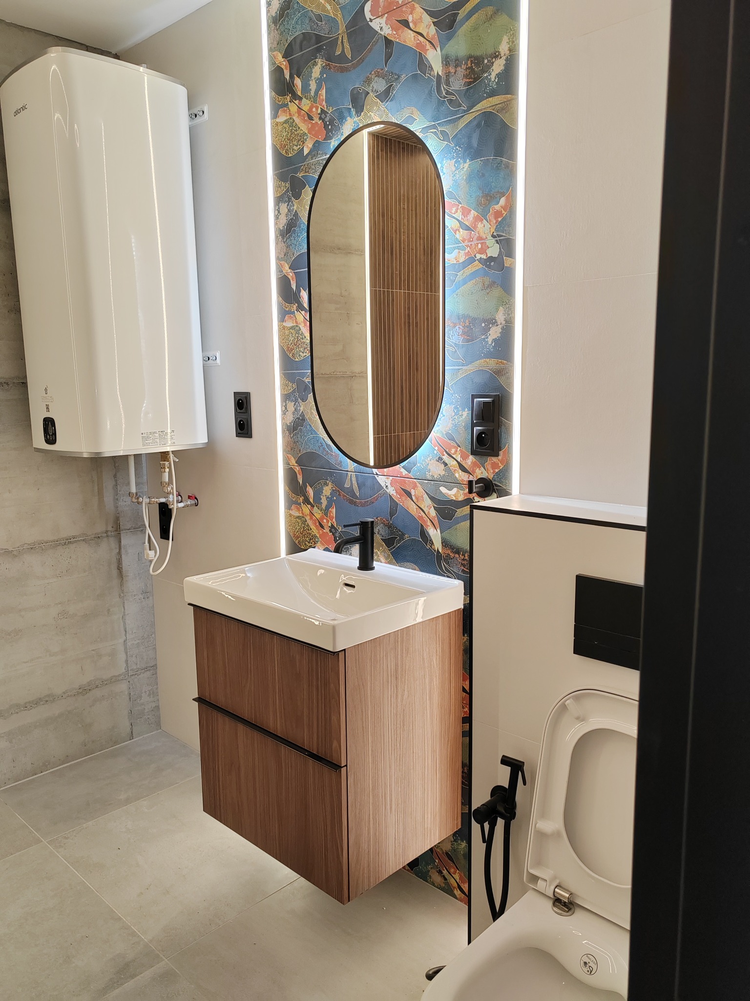 Nowoczesna łazienka z owalnym lustrem podświetlonym LED, umywalką z szafką z drewnopodobnym fornirem, wiszącym bojlerem Atlantic i toaletą z bidetką w kolorze czarnym, na tle płytek imitujących...