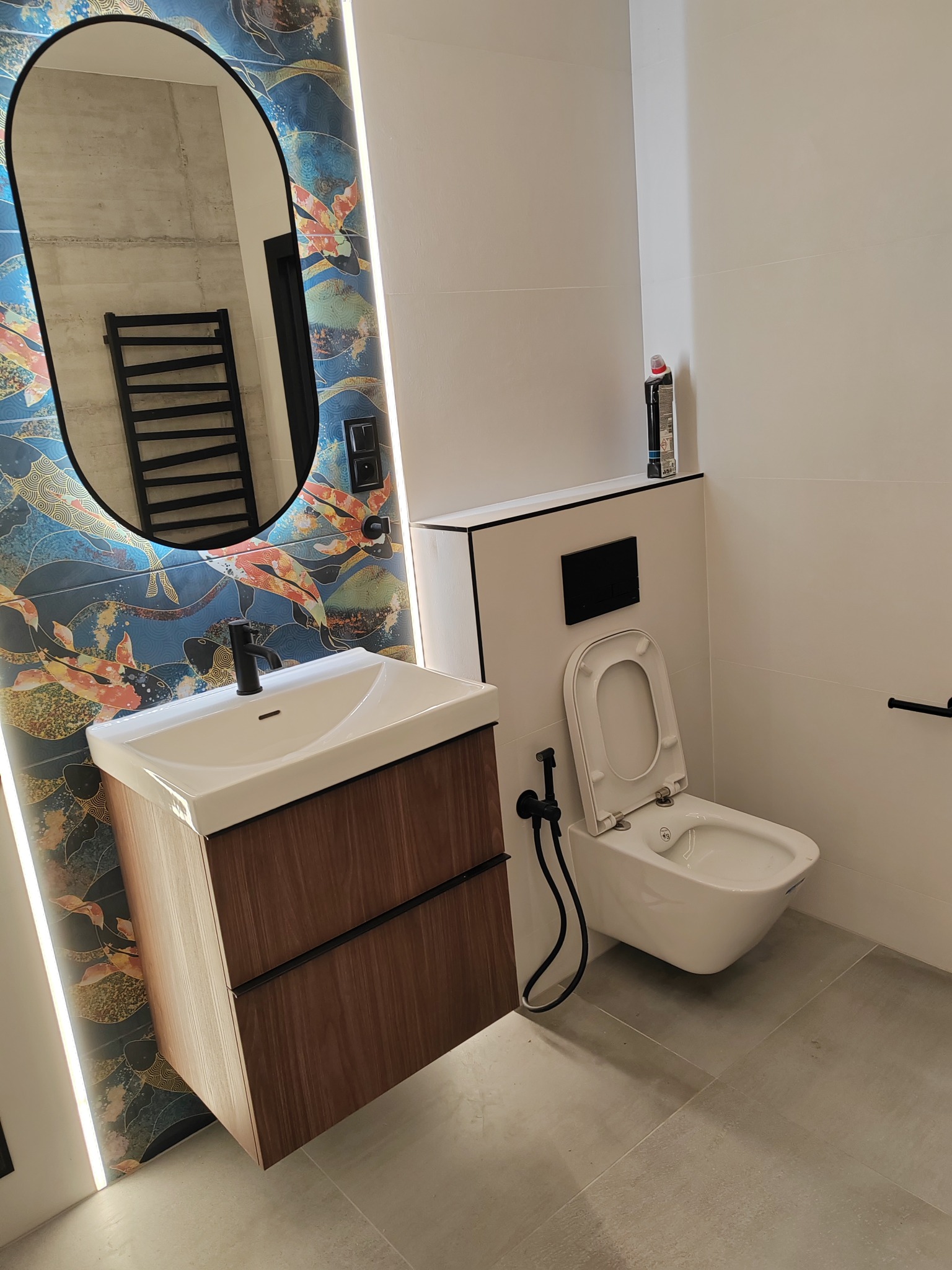 Nowoczesna łazienka z umywalką na drewnianej szafce, toaletą podwieszaną z bidetką i owalnym lustrem podświetlonym LED na ścianie z tapetą w orientalne wzory.