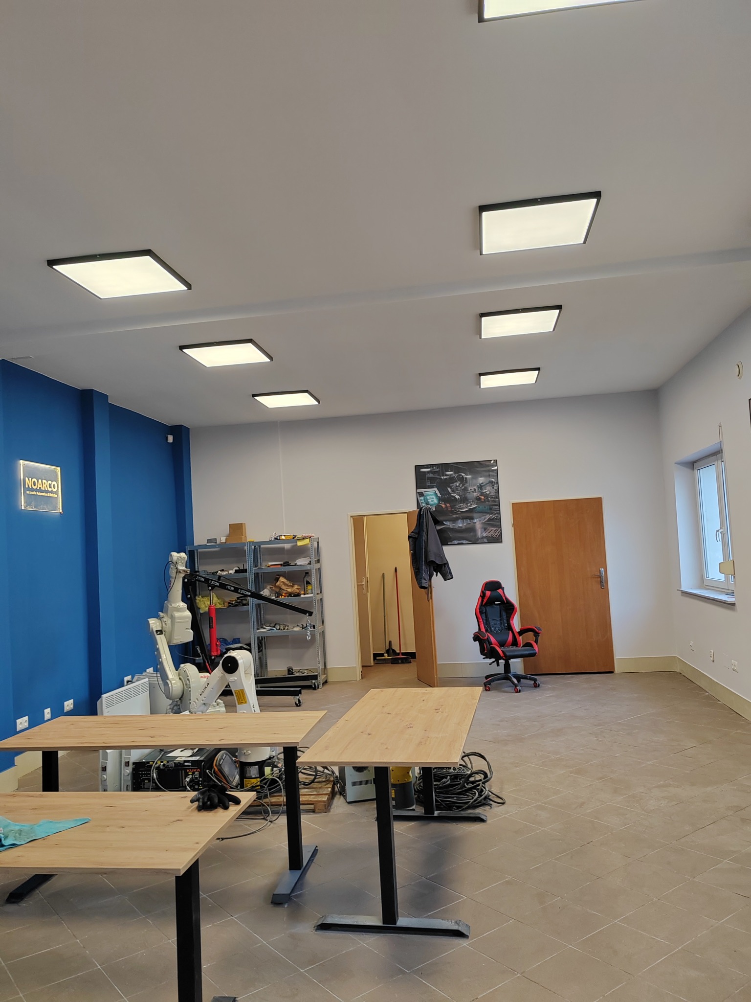 Przestronne, jasne wnętrze biura z niebieską ścianą, wyposażone w robotyczne ramię KUKA, biurka z czarnymi nogami, krzesło gamingowe i równomiernie rozmieszczone kwadratowe lampy sufitowe.