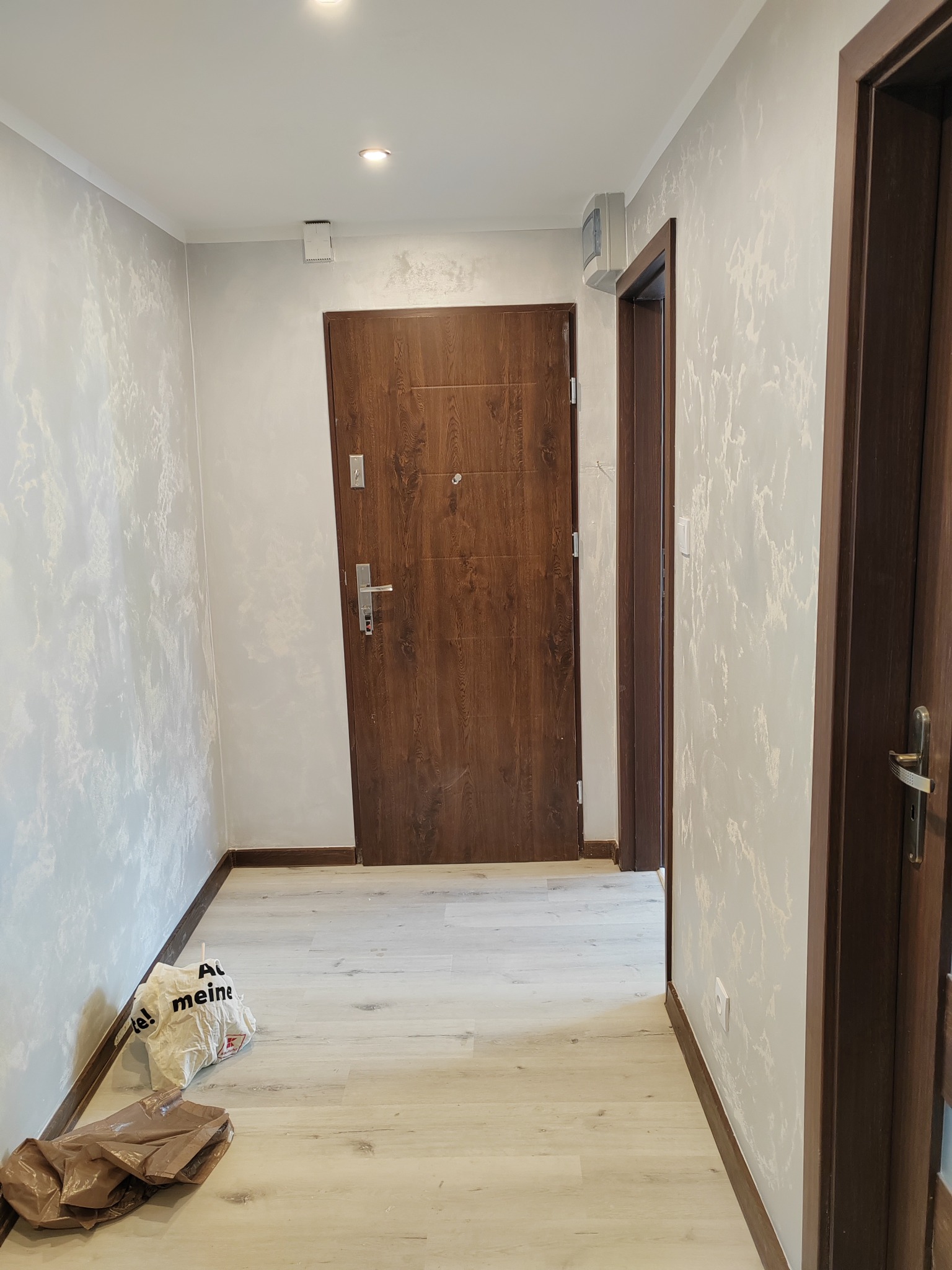 Korytarz po remoncie z drewnianymi drzwiami, panelami podłogowymi i ścianami z efektem dekoracyjnym, widoczne torby po zakupach przy ścianie.