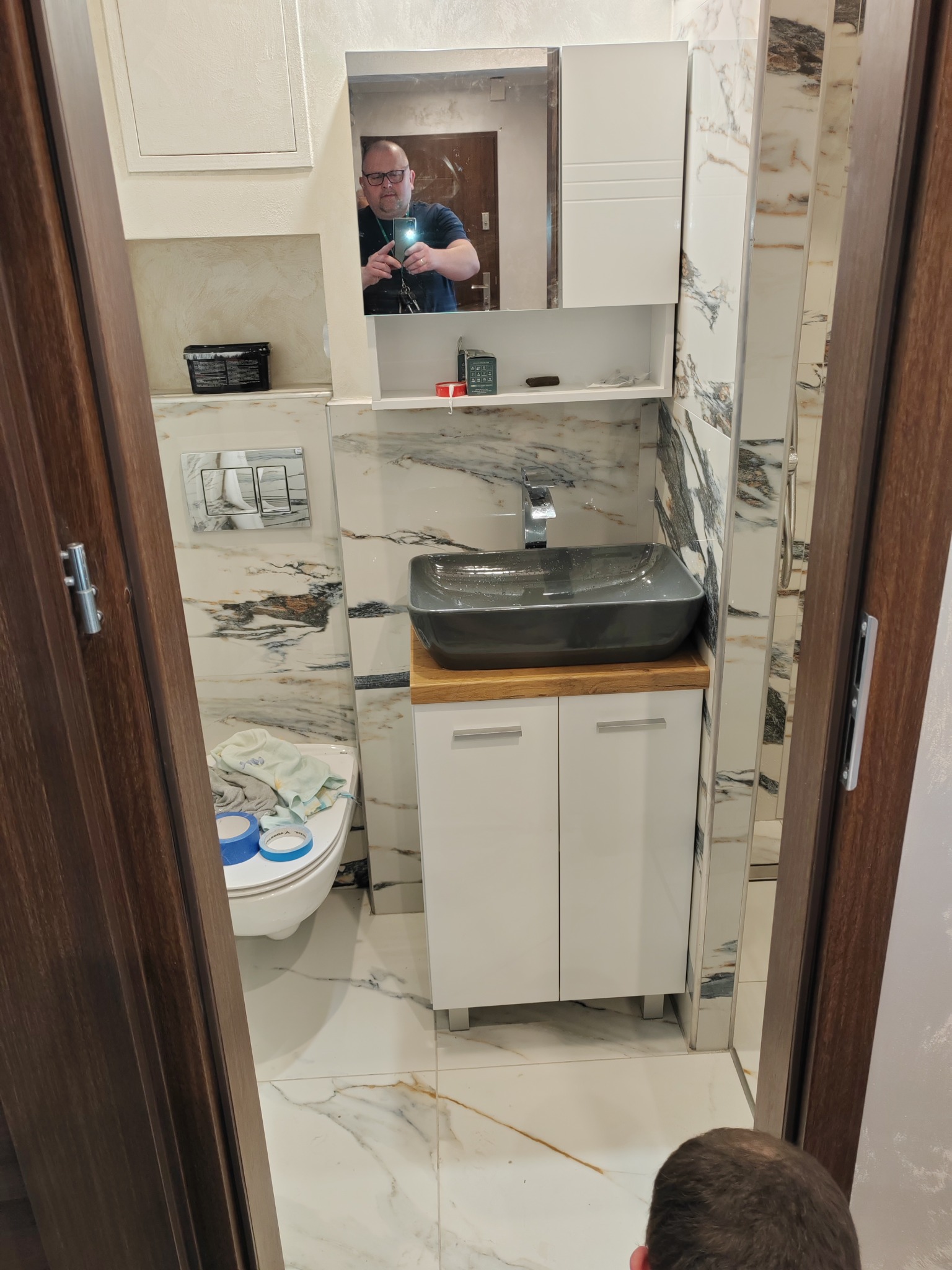 Wykończona łazienka z marmurowymi płytkami, szarą umywalką na drewnianym blacie i białymi szafkami pod umywalką, widok przez futrynę drzwi z odbiciem fotografa w lustrze.