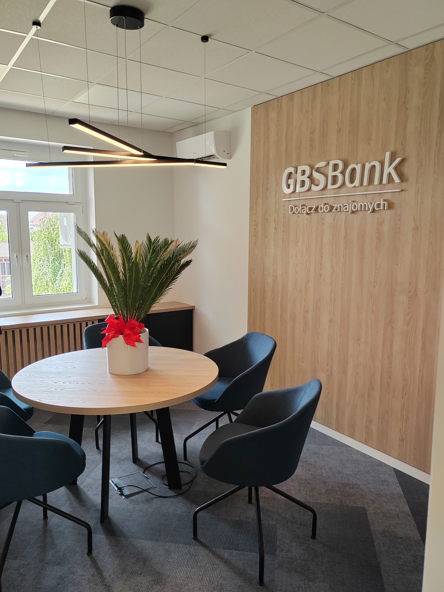 Jasne biuro z okrągłym stołem, czterema granatowymi krzesłami, rośliną w białej doniczce z czerwoną kokardą, nowoczesną lampą sufitową i ścianą z drewnianym panelem z logo GBS Bank.