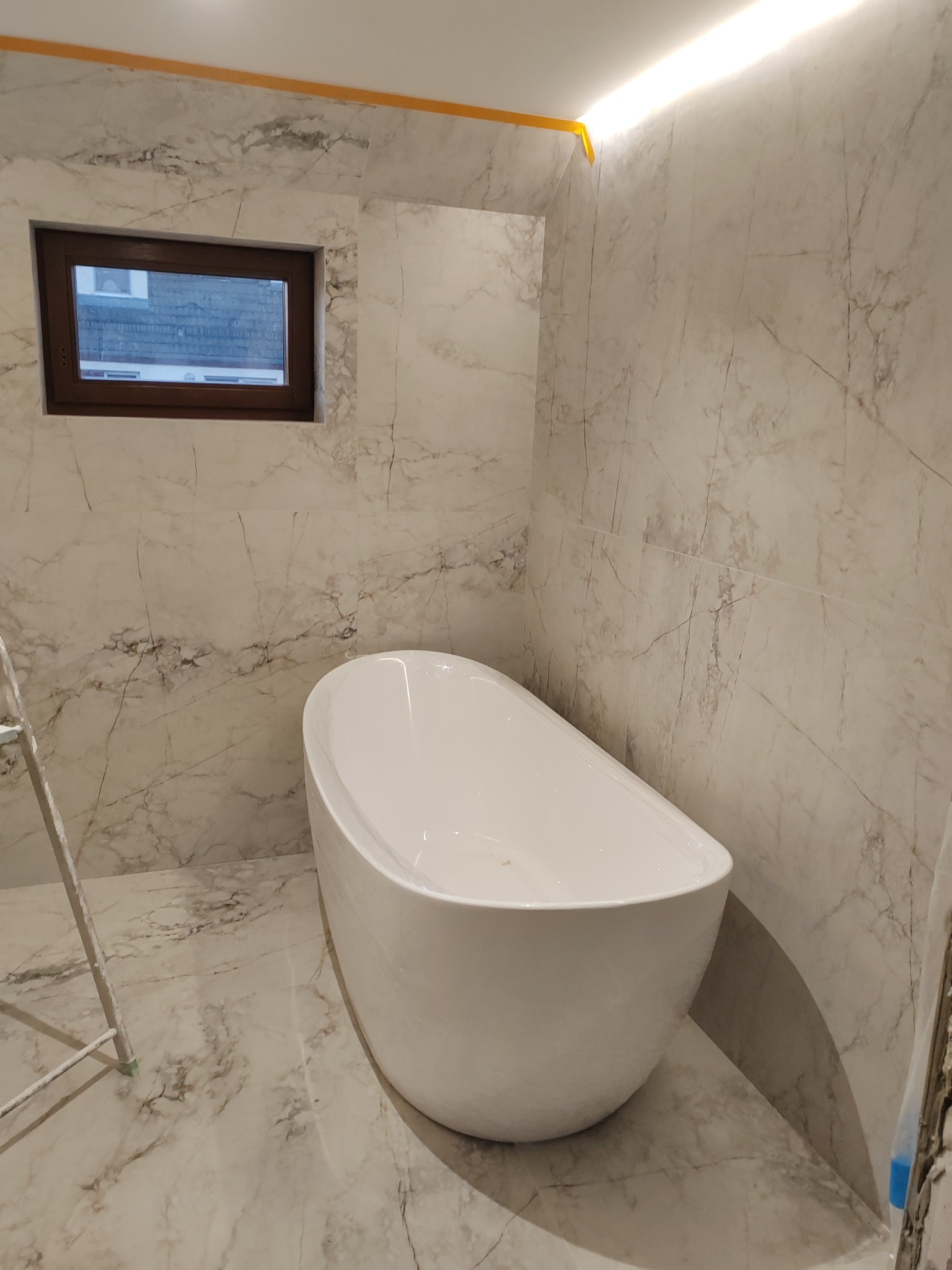 Nowoczesna, wolnostojąca wanna w łazience z marmurowymi płytkami, oświetleniem LED podsufitowym i małym oknem z brązową ramą.