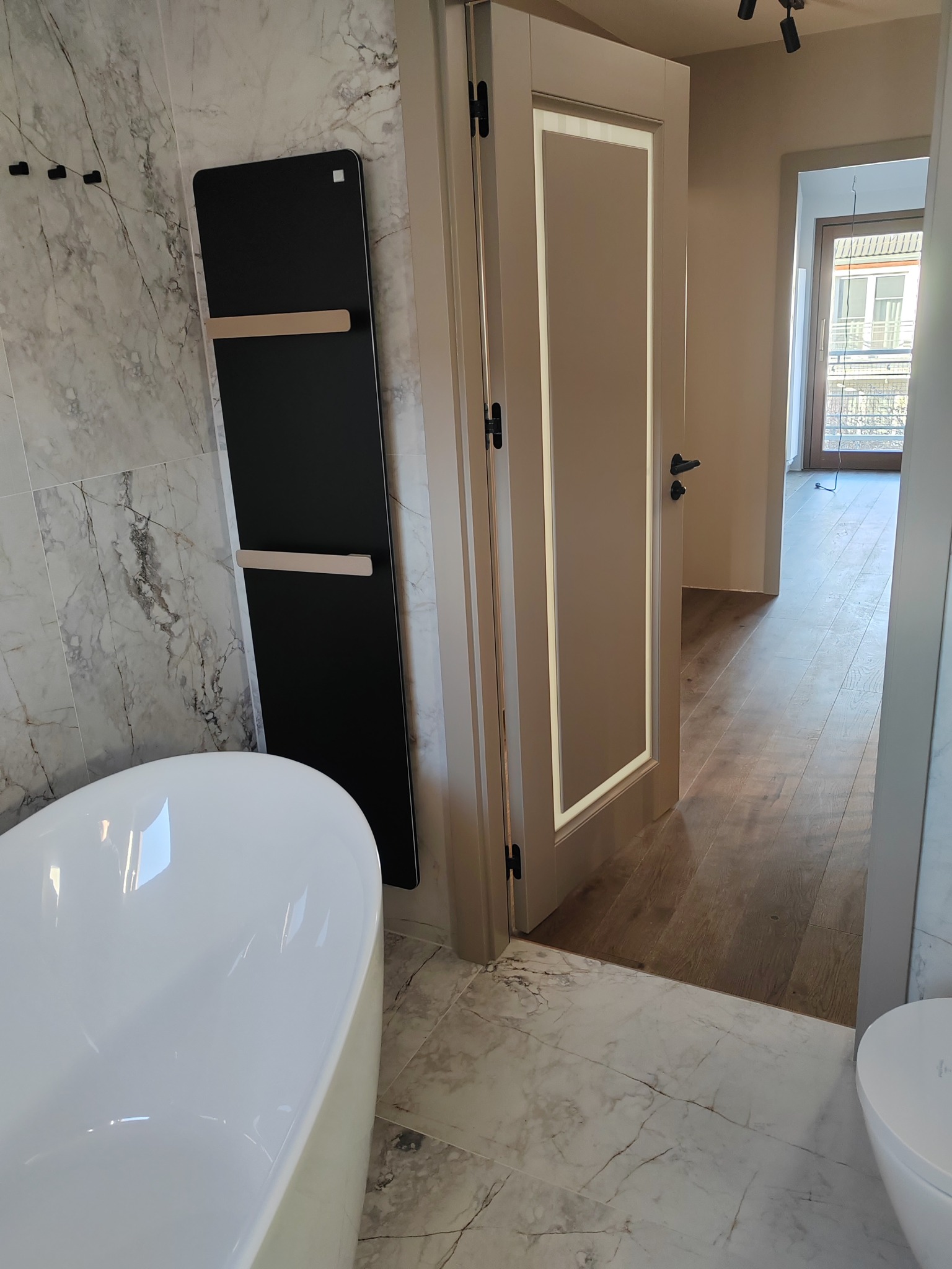 Nowoczesna łazienka z marmurowymi płytkami, białą wanną, czarnym grzejnikiem drabinkowym i oświetloną wewnętrznie ramą drzwi.