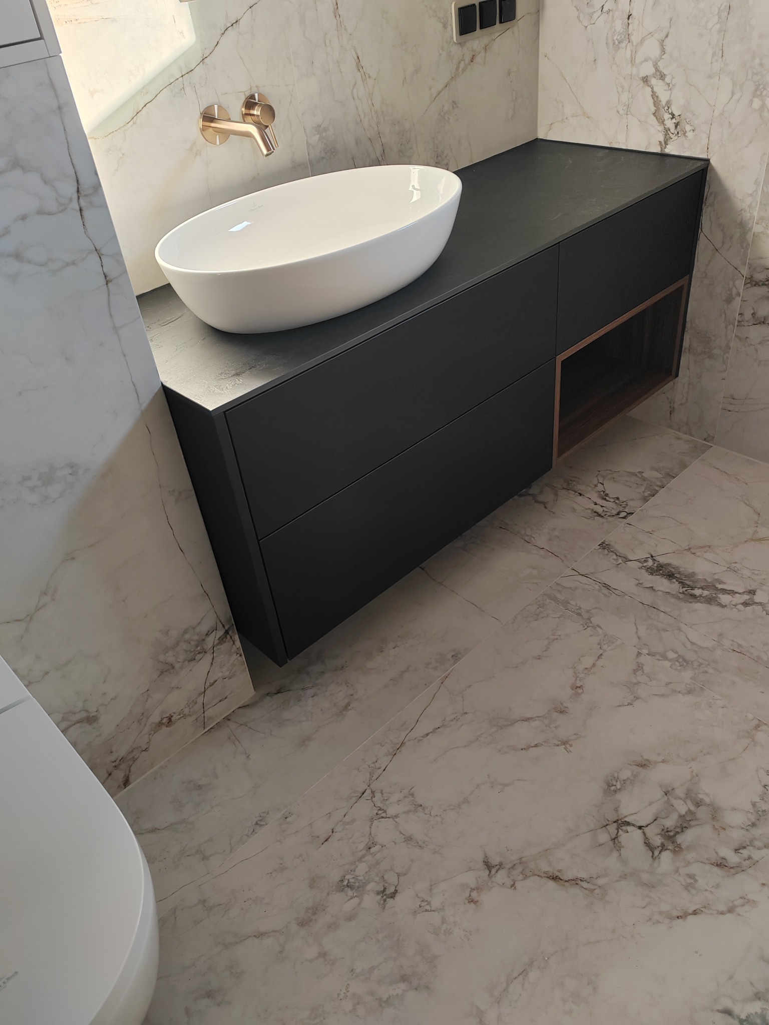 Nowoczesna łazienka z białą, owalną umywalką nablatową, czarną szafką pod umywalką z otwartą półką, złotą armaturą i marmurowymi płytkami na ścianach i podłodze.