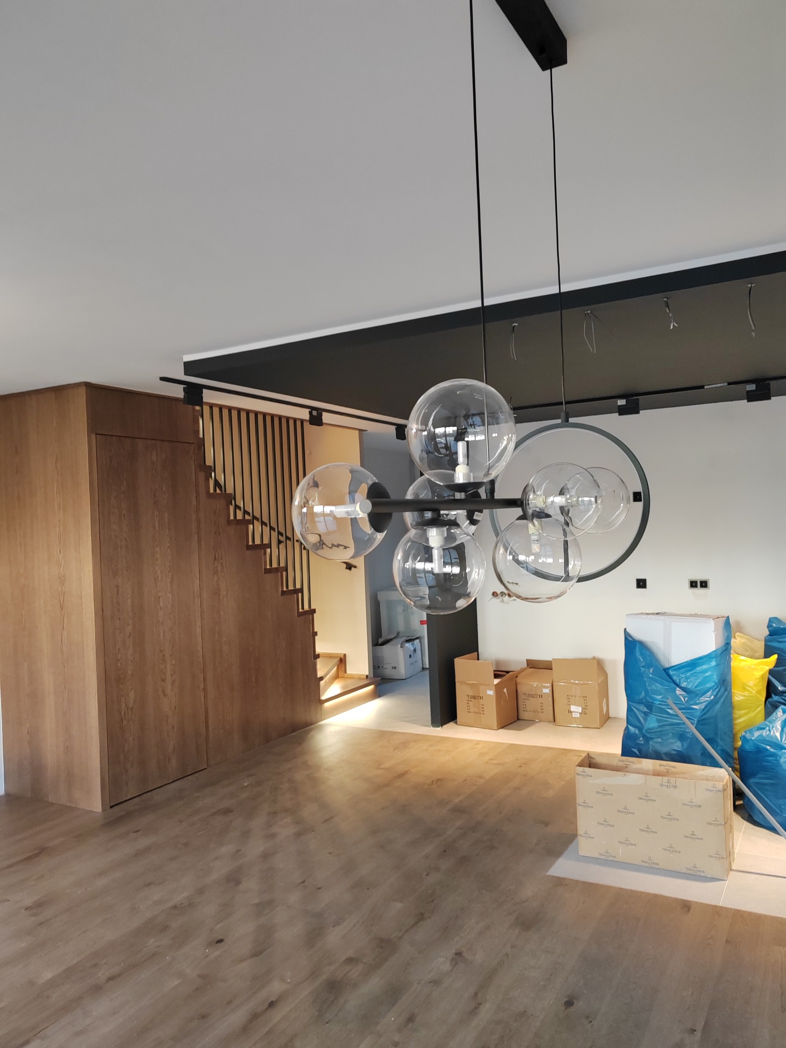 Nowoczesny żyrandol z kulistymi kloszami w jasnym wnętrzu z drewnianą podłogą i schodami, widoczne kartony i worki budowlane sugerujące prace wykończeniowe.